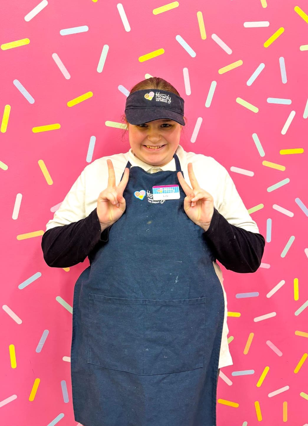 Lilly, a Howdy Homemade Hero posing for a photo at work, holding up peace signs, with a big smile.